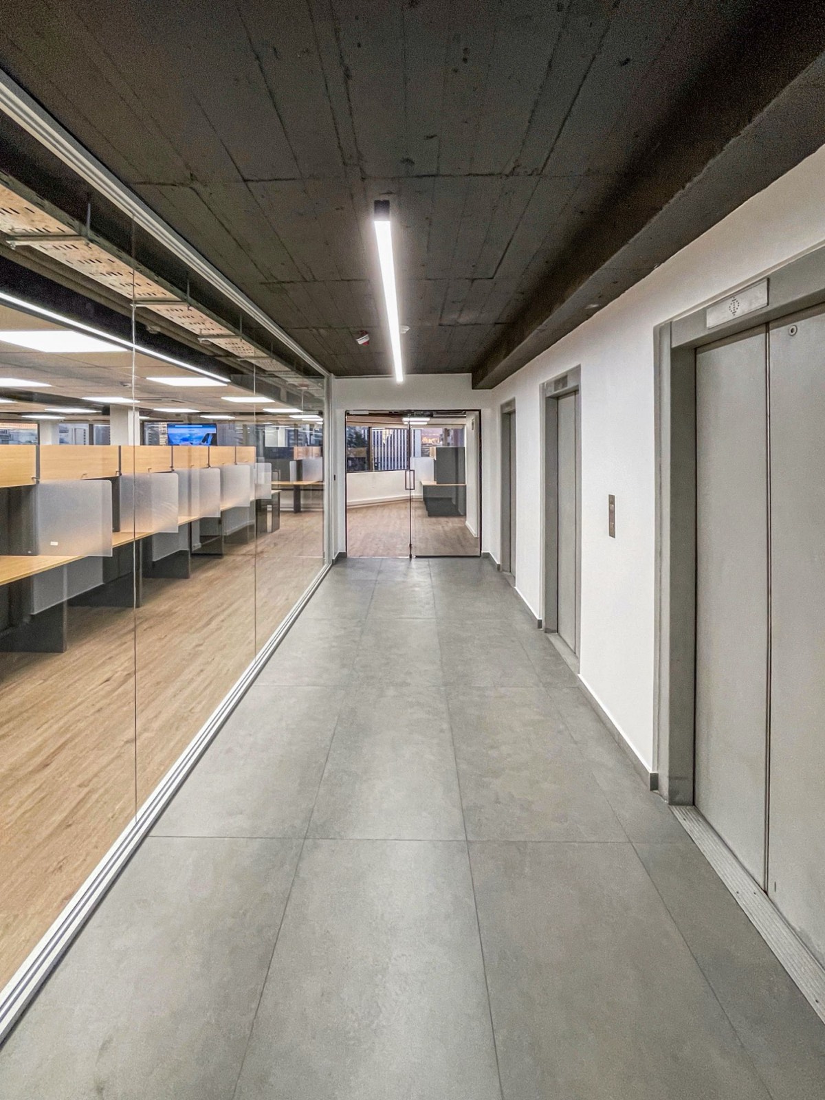 Hall de ascensores con piso gris y vista a los puestos de trabajo tras el muro de vidrio